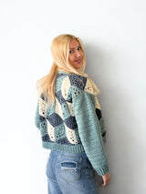 Back view of woman standing in front of a white brick wall. She wears a crochet cardigan in blue-green, dark denim, and cream. The sleeves are solid and the back has a 3-dimensional design made of granny motifs. thumbnail