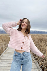 Woman stands with arms out on wooden boardwalk, wearing fuzzy pink crochet cardigan. thumbnail