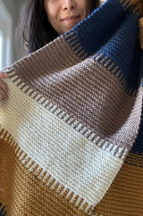 Woman holds knit wrap toward camera, showing garter textures and bands of vertical stripes in cream, taupe, blue, and golden yellow. thumbnail