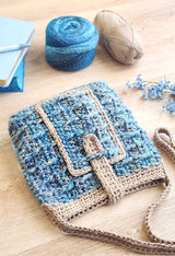 Textured crochet bag laying on wooden table, opening facing the camera. Yarn balls and a stock of journals in the background. thumbnail