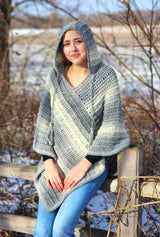 Woman sits on wooden fence in front of snowy field, wearing crochet poncho with hood worn up. thumbnail