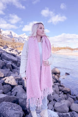 Woman stands on rocky shoreline with mountains in the distance. She wears jeans, a cream coat, and a long pink fluffy scarf. The scarf reaches below her knees. thumbnail