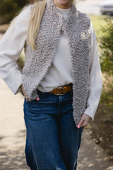 Woman in jeans, brown belt, and a white top stands on a sidewalk. She has a knit tan vest on. The vest reaches her lower hip and is open at front, with a standing collar. thumbnail