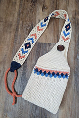 Flat lay of a crochet sling bag. The main bag is cream and has a big button closure. The strap has a geometric pattern in navy, blue, and orange. There is an orange cord to adjust the strap length. thumbnail