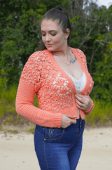 Woman stands on a sandy road, with greenery in the background. She wears a white camisole and blue jeans, and a coral lace crochet cardigan. thumbnail