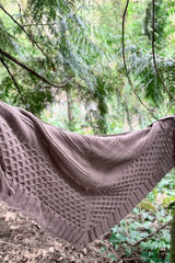 Tan cabled shawl draped over a branch in a conifer forest. thumbnail