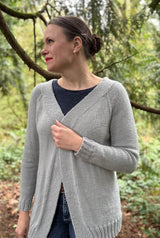 Front view of pale grey cardigan, showing wide front bands, long sleeves, and raglan lines. Woman wears it over a dark top and jeans, standing in a conifer forest. thumbnail