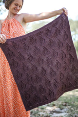 Woman wearing an orange summer dress holds a square baby blanket out to the side. The blanket ruffles in the breeze, it is purple-brown with an all over textured pattern. thumbnail