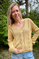 Woman leans to the side while wearing a golden yellow sweater and blue jeans, standing in front of some greening bushes. The sweater has three rope cables running down the front, with one centered on the v-neck opening. thumbnail