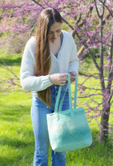 Woman in cream sweater and jeans stands outdoors in front of flowering tree. She holds a green crochet toe bag with long shoulder straps. thumbnail