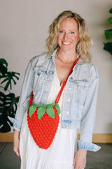 Woman in pale denim jacket and white dress wears strawberry bag crossbody style. thumbnail