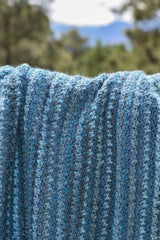 Close up of crochet blanket in shades of blue with trees and a mountain in the distance. thumbnail