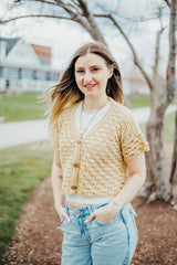 Woman in a pale yellow short sleeve cardigan over a white t-shirt, with pale blue jeans. She stands in a mulches area in front of a bare tree, with a house in the background. thumbnail