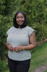 Front view of woman wearing pale blue-green knit tee and dark pants. She stands with her hands clasped at the waist, in front of an evergreen hedge. thumbnail