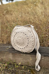Pale beige crochet bag sits on a railroad tie in a field of golden grass. The long strap of the bag hangs to the side, and the top part of the circular body is open. thumbnail