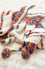 Close up on pompoms on corner of crochet blanket. The poms and some stripes in the blanket are multi-color in amber, pink, purple, and blue. The other stripes in the blanket are white. thumbnail