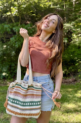 Smiling woman stands in a backyard in front of a treeline. She holds a crochet tote bag in the crook of her elbow. The bag is cream with stripes of amber, sage green, and pale pink. thumbnail