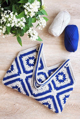 Bag laying on wooden surface with handles folded over bag body. There is a potted plant with green leaves and white flowers in upper left corner, and two balls of yarn in upper right. thumbnail
