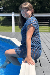 Side view of woman looking at camera while sitting on a pool deck, with legs in the water. She wears a sleeveless crochet mesh coverup that goes to mid-thigh. thumbnail