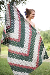 Front view of woman holding crochet blanket up towards camera. It has a chevron design with stripes of varied width. thumbnail