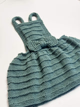 Flat lay of knit pinafore dress on white background. The skirt and bodice have a ridged texture and a bow at the waist. thumbnail
