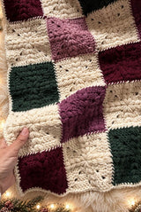 Top down view of crochet blanket showing seams between squares and border. A hand in the lower left corner holds blanket edge above white furry background with twinkle lights. thumbnail
