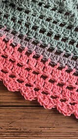 closeup view of the textured stitch pattern and lacy border on a multicolored pastel pink, grey, and blue gradient crocheted triangle shawl resting on a wooden surface thumbnail