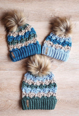three handmade striped crochet beanies with faux fur pom-poms and ribbed cuffs arranged on a light wooden surface thumbnail