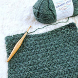 close-up of forest green crocheted scarf in sedge stitch with wooden crochet hook and skein of Lion Brand Heartland yarn thumbnail
