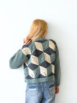 Back view of woman standing in front of a white brick wall. She wears a crochet cardigan in blue-green, dark denim, and cream. The sleeves are solid and the back has a 3-dimensional design made of granny motifs.  thumbnail
