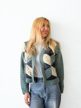 Front view of woman wearing crochet cardigan with white top and pale jeans, standing in front of white brick wall. The cardigan is blue-green, dark denim blue, and cream.  thumbnail