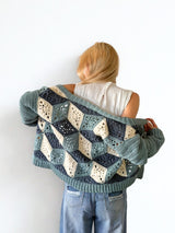 Back view of woman standing in front of a white brick wall. She wears a crochet cardigan in blue-green, dark denim, and cream. The sleeves are solid and the back has a 3-dimensional design made of granny motifs.  thumbnail