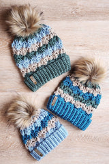 three handmade striped crochet beanies with faux fur pom-poms and ribbed cuffs arranged on a light wooden surface thumbnail
