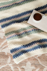 striped crocheted blanket with blue, sage green, and beige stripes laid out on a beige patterned rug with a white coffee mug on a saucer beside it thumbnail