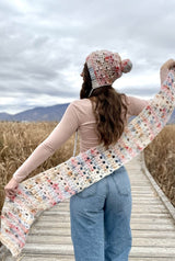 Woman holding scarf behind back, facing away from camera, while wearing bonnet with pompom. thumbnail
