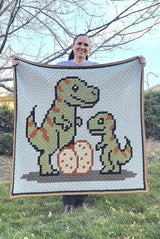 Woman holds up crochet afghan with t-rex family while standing outdoors.  thumbnail