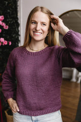 Woman wears purple sweater indoors, in front of wall of greenery thumbnail
