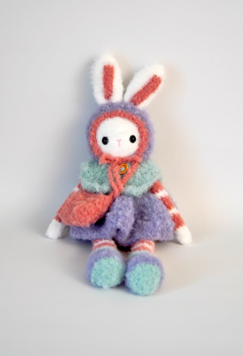 Fuzzy crochet bunny with purple skirt, red & white striped arms & legs, and aqua vest. sitting up against white backdrop. 