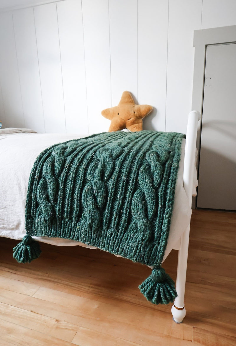 Green cabled throw on a white bed, with a golden star pillow in background. 