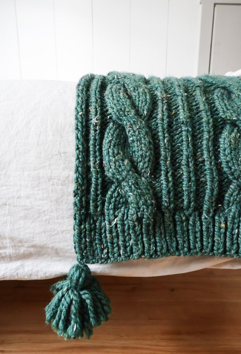 Close up of green blanket hem, with large cable, ribbing and tassel.