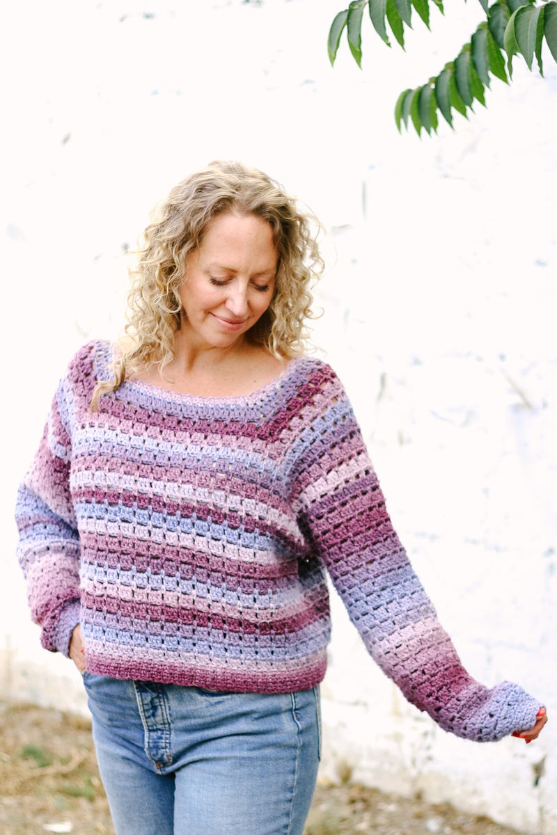 Woman with blonde curly hair wears pink & purple crochet sweater with blue jeans. 
