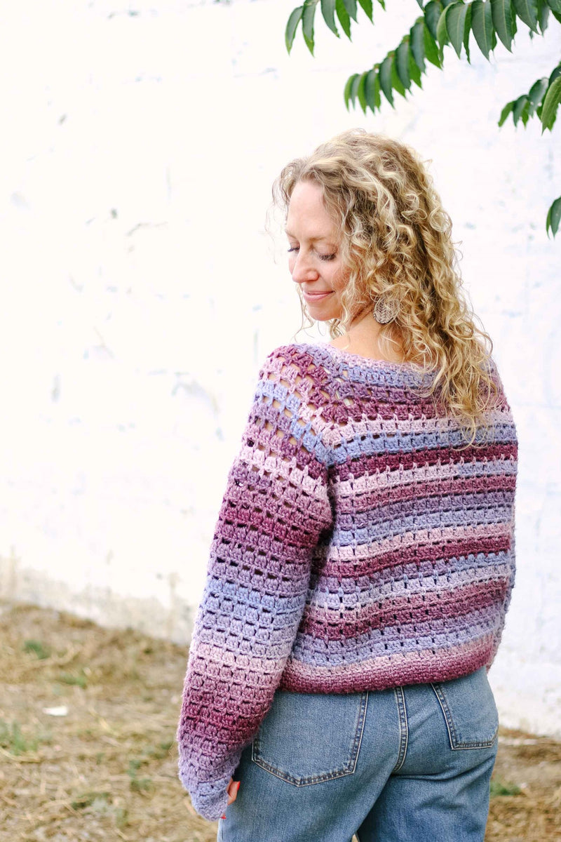 Back view of purple & pink crochet sweater worn by model with curly hair and blue jeans. 