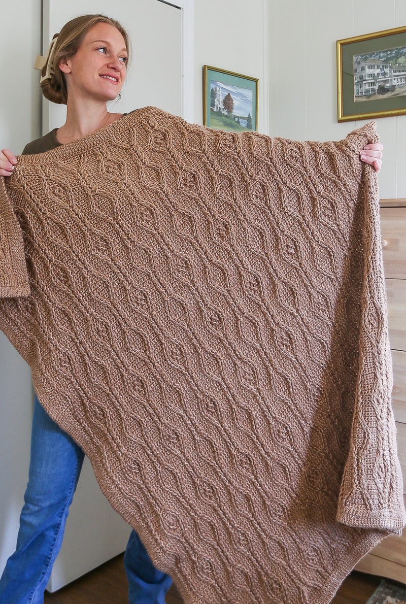 Woman holds tan crochet blanket, covering her from shoulder to ankle. 