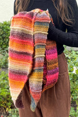Woman wearing black turtleneck and brown pants holds multicolor crochet shawl in front of her.  thumbnail