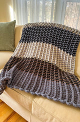 Blanket with striped color block sections in shades of purple, tan, and blue on pale couch with green throw pillow.  thumbnail