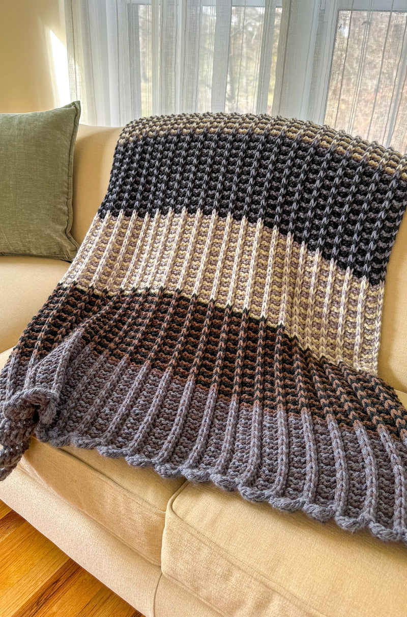 Blanket with striped color block sections in shades of purple, tan, and blue on pale couch with green throw pillow. 