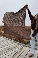 Brown afghan with mesh design held like a banner by woman on a boardwalk in a wetland.  thumbnail
