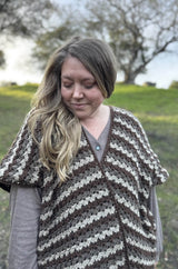 Woman with blonde hair wears striped crochet cape in shades of cream and brown, standing outside.  thumbnail
