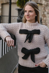 Woman wears tan crochet sweater with two black bows on front, leaning against fence.  thumbnail