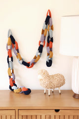 Paper chain style crochet garland in dark navy, orange, lavender, mustard, and aqua hangs on cream wall above wood sideboard.  thumbnail